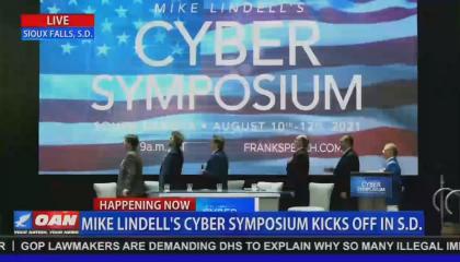 Screenshot of an OAN broadcast of MyPillow CEO Mike Lindell and a panel pledging allegiance to a large American flag video, mid-transition from a screen reading "Mike Lindell's Cyber Symposium." OAN chyron reads "Mike Lindell's Cyber Symposium Kicks Off In S.D."
